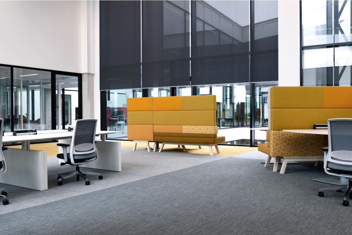 An open office space with modern furniture, including yellow and gray sectioned seating areas, and large windows with Crestron motorized window treatments.
