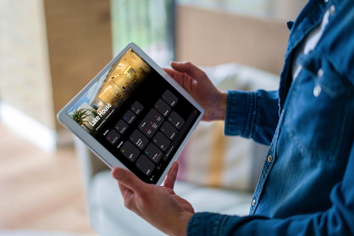 Person holding a tablet displaying Crestron home automation controls for "Lake House" with various room options and settings.