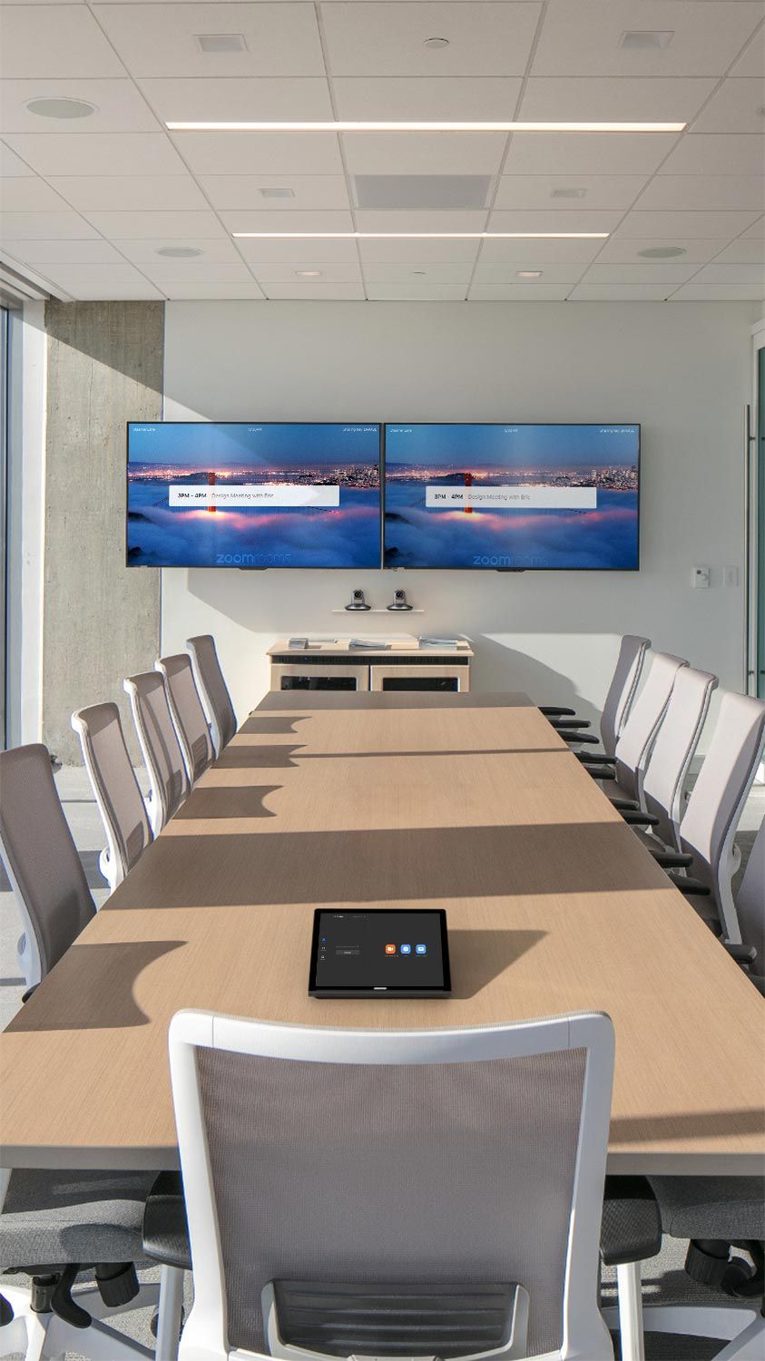 A modern conference room with a long wooden table, white office chairs, and dual screens displaying a Crestron meeting interface.