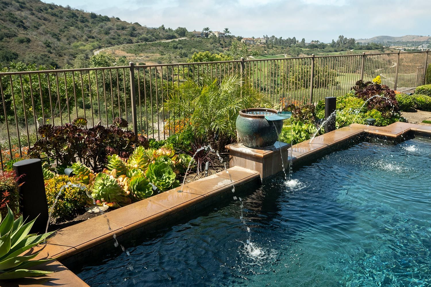 Pool with water fountains and James Loudspeaker outdoor speakers, surrounded by lush greenery and a scenic hilly background.